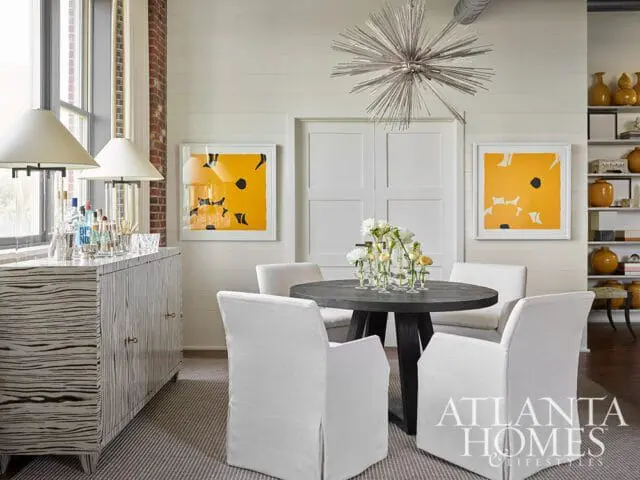 Dining area with a dark wood table, four white slipcovered chairs, a striped sideboard with a bar setup, and abstract yellow artwork on a shiplap wall.