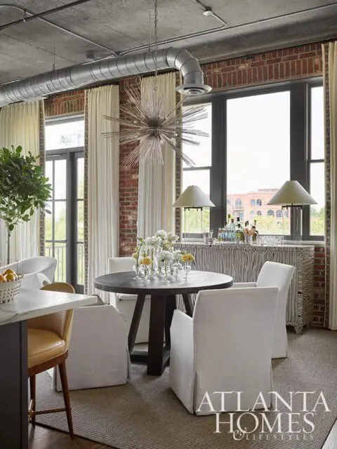 Industrial-style dining area with exposed brick walls, concrete ceiling, and ductwork. Features a dark wood round dining table with white slipcovered chairs, a Sputnik chandelier, and a textured sideboard with lamps.