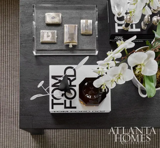 Dark wood coffee table with a Tom Ford book, decorative objects, and white orchids. Atlanta Homes & Lifestyles logo in the bottom right corner.