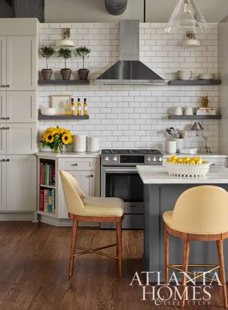 Gray and white kitchen with white subway tile backsplash, stainless steel range hood, light gray cabinetry, and two yellow upholstered bar stools at a dark gray island.