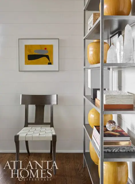 Modern living room with shiplap walls, a dark wood chair with white woven seat, abstract art, and a metal shelving unit displaying yellow vases and crystal objects.