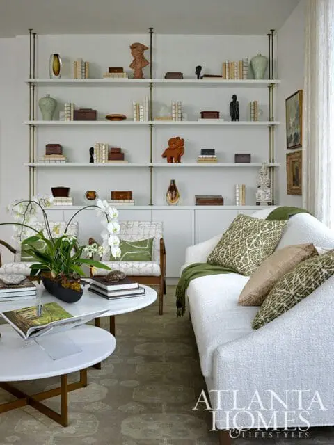 Living room with white textured sofa, green patterned pillows, white marble coffee tables, and a large white bookshelf filled with books and decorative objects.
