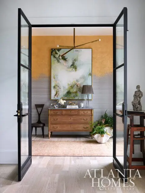View through black-framed glass doors into a room with a wooden dresser, abstract art, and a modern chandelier. The wall features a gold and grey textured finish.
