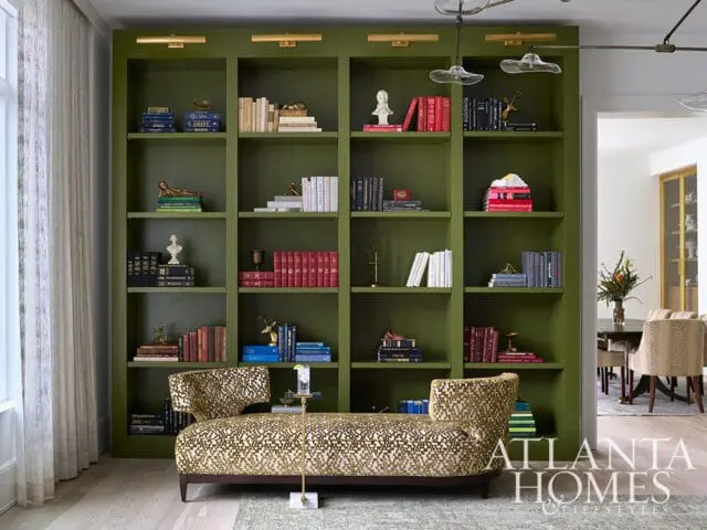 Living room with a large olive green built-in bookshelf filled with books and decorative objects. A patterned chaise lounge sits in front of the bookshelf.