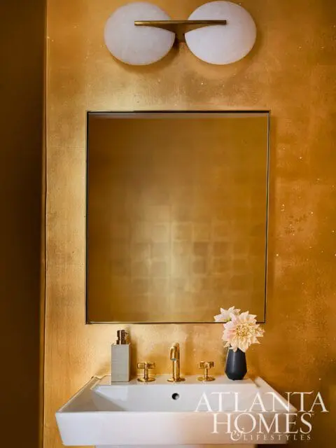 Powder room with gold textured walls, a square mirror, brass fixtures, a white sink, and a modern wall sconce with two white globes.