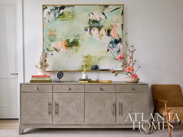 Light gray wood sideboard with brass hardware topped with books, a bust, and a floral arrangement, beneath a large abstract painting.
