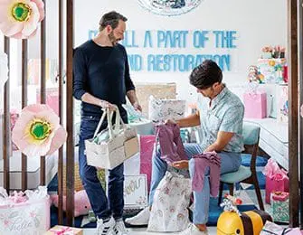 Two men in a room decorated for a baby shower, surrounded by gifts and pastel-colored decorations. One man holds a gift basket, the other examines baby clothes.