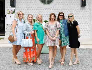 Six women in dresses and casual wear pose for a photo in front of a white textured building with a circular window.