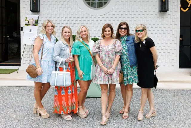 Six women in dresses and casual wear pose for a photo in front of a white textured building with a circular window.