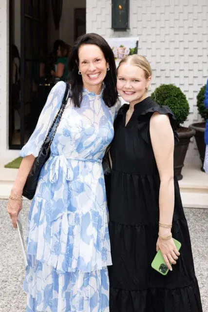Two women smiling at the camera outside a white brick house. The woman on the left wears a blue and white floral dress and the woman on the right wears a black dress.