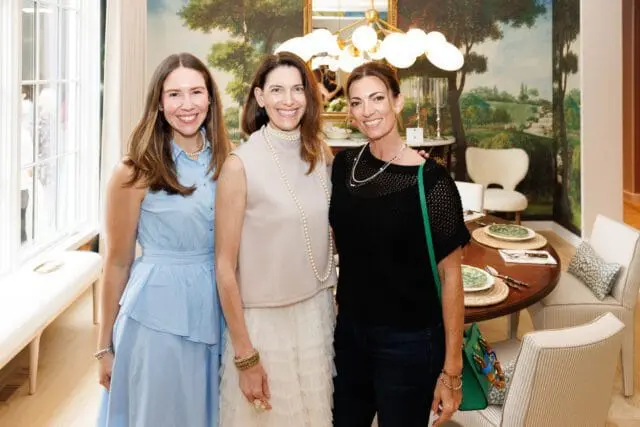 Three women posing in a dining room with a large mural, a round table set for dinner, and striped upholstered chairs.