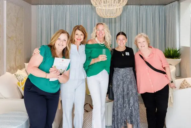 Five women pose for a photo in a room with light blue patterned curtains, a large woven pendant light, and white furniture. One woman holds a brochure for Southeastern Showhouse.
