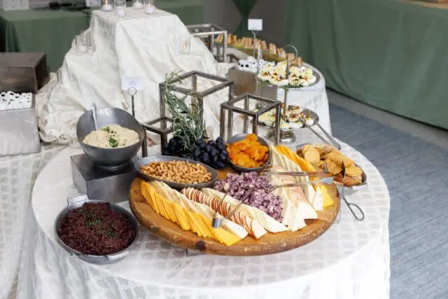 Buffet table with a round wooden platter of sliced cheeses, crackers, grapes, dried apricots, and nuts. Other dishes include deviled eggs, hummus, and a dark spread.