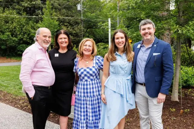 Five people pose for a photo outdoors. From left to right: a man in a pink button-down shirt, a woman in a black dress, a woman in a blue and white gingham dress, a woman in a light blue dress, and a man in a blue blazer over a blue and white checkered shirt.