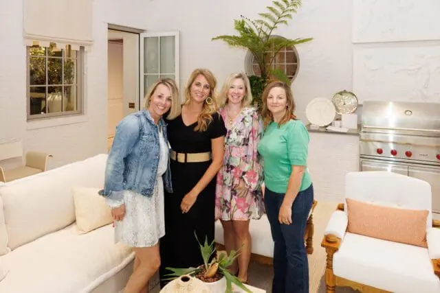 Four women pose in a bright, modern living space featuring a white textured sofa, a built-in Wolf grill, and white brick walls with decorative reliefs.
