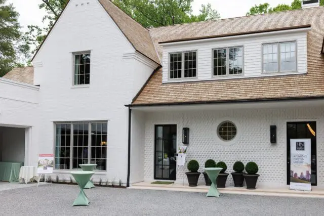 White brick house with cedar shake roof, large windows, and a black front door. Outdoor cocktail tables and potted topiaries are set up in front.