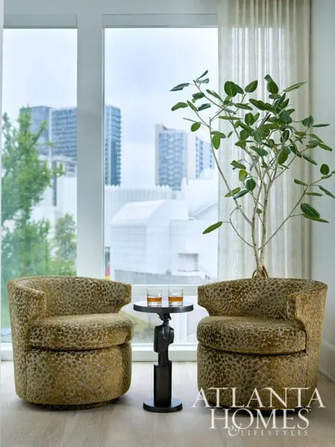 Two upholstered swivel chairs with a leopard-like pattern flank a small dark metal table holding two glasses of amber liquid. A tall plant stands behind the table.
