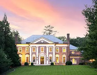 Neoclassical brick mansion with white columns and a pedimented portico, set on a manicured lawn with mature trees under a pastel sunset sky.