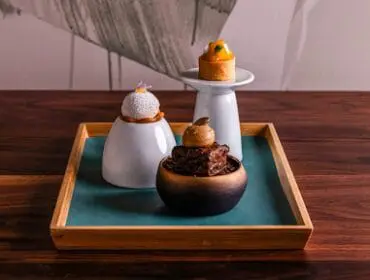 Three artfully presented desserts on a wooden tray with a teal mat. One dessert is a white sphere topped with a purple flower, another is a dark chocolate square with a caramel topping, and the third is a small tart with orange fruit.