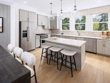 Light gray kitchen with a marble island, stainless steel appliances, light wood floors, and a dining table with white upholstered chairs.