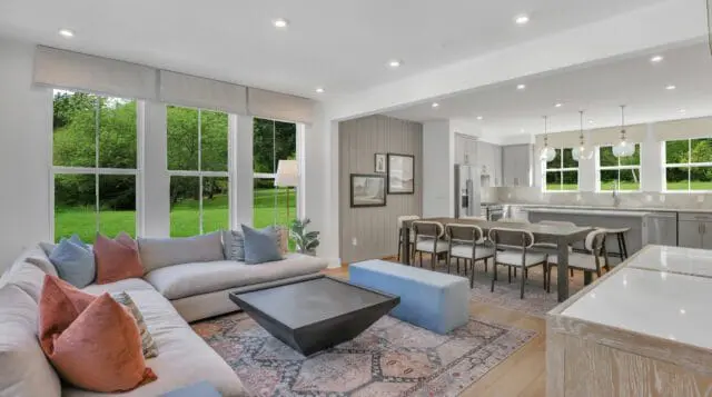 Open concept living and dining area with a sectional sofa, geometric coffee table, dining table with chairs, and a light gray kitchen with island. Large windows overlook a lush green lawn and trees.