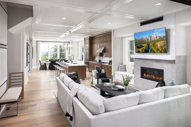 Open-concept living and dining area with white sofas, a dark wood kitchen with stainless steel appliances, and a fireplace with a TV above.