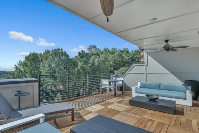 Modern balcony with wooden plank flooring, light blue sofa, dark coffee table, and lounge chairs, overlooking a tree-filled landscape with a building on a hill.