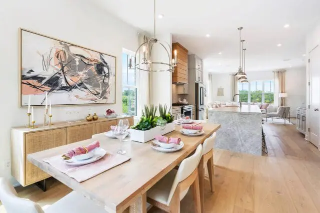 Dining room with light wood table set with pink napkins and white plates, adjacent to a kitchen with a marble island and stainless steel appliances.