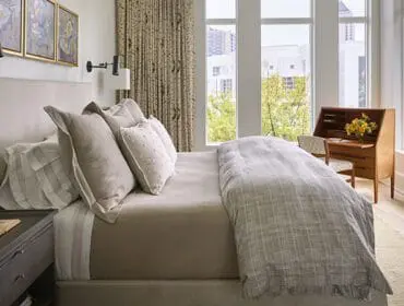 Bedroom with a king-size bed featuring a neutral upholstered headboard, layered pillows, and a patterned duvet. A wooden secretary desk with a floral arrangement sits by the window.