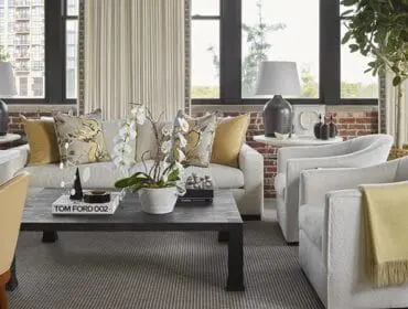 Living room with white sofa, two white armchairs with yellow throw, dark coffee table with books, and large windows overlooking a city building.