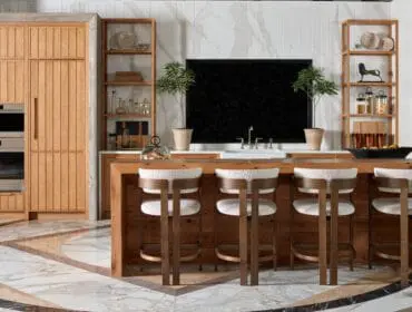 Modern kitchen with light wood cabinetry, a large wood island with white boucle bar stools, marble floors with geometric inlay, and a black backsplash.