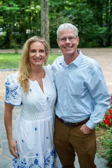 A smiling couple poses for a photo outdoors, with the woman wearing a white dress with blue embroidery and the man in a light blue button-down shirt and brown pants.