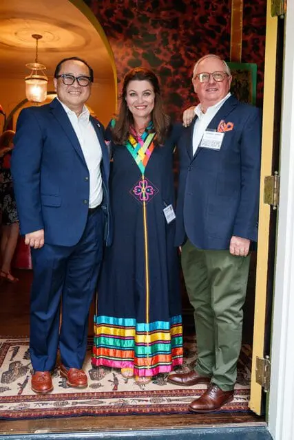 Three people posing for a photo. A man in a navy suit and glasses stands on the left. A woman in a dark dress with colorful trim stands in the middle. A man in a navy blazer and glasses stands on the right.