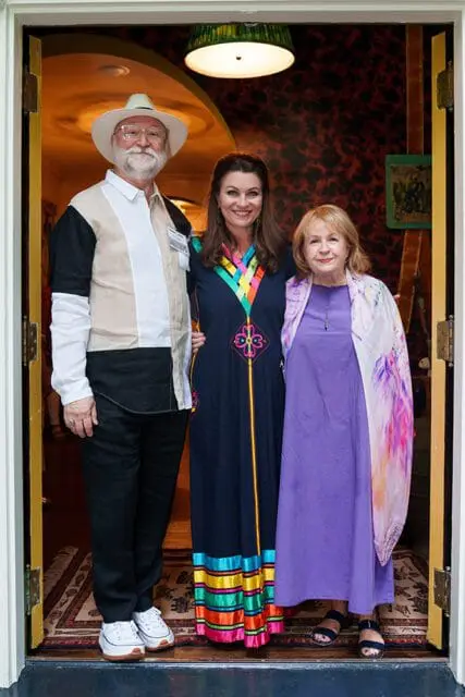 Man in a white hat and color-blocked shirt, a woman in a dark blue dress with colorful trim, and a woman in a purple dress with a patterned shawl stand together in a doorway.