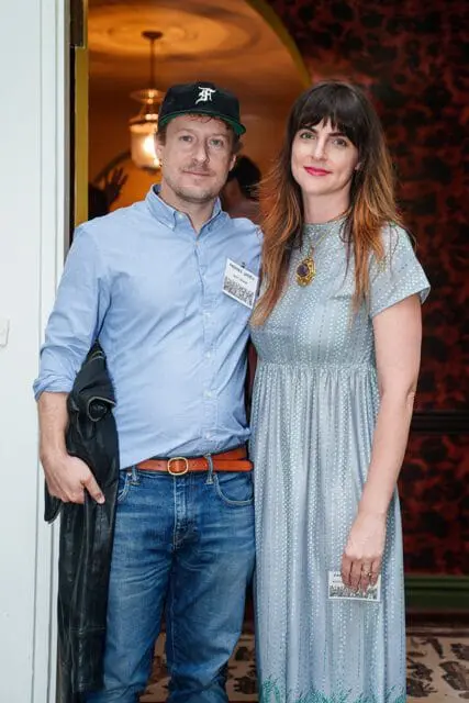 Man in blue button-down shirt and jeans wearing a black baseball cap, and woman in a light blue patterned dress wearing a pendant necklace. Both are wearing name tags.
