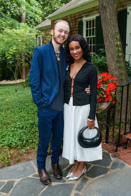 A smiling man in a blue suit and a woman in a black top and white skirt stand together outdoors in front of a house.