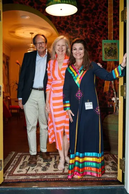 Three people stand on a patterned rug in a doorway. A man in a navy blazer and tan pants stands on the left. A woman in a pink and orange striped dress stands in the middle. A woman in a navy dress with colorful trim stands on the right.