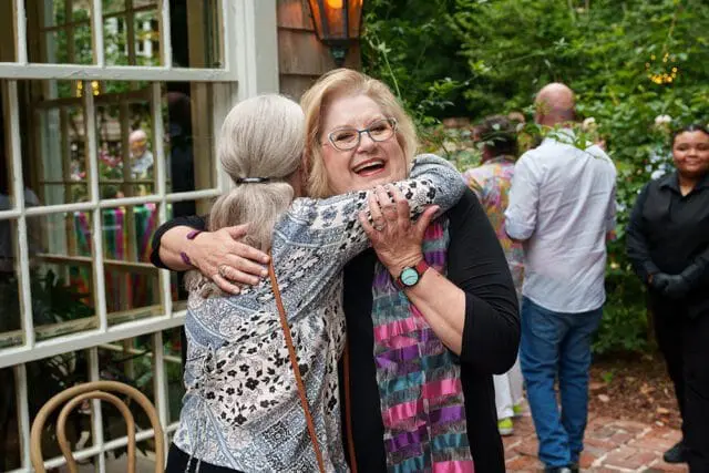 Two women embracing outdoors, one wearing glasses and a colorful scarf, with other guests in the background.