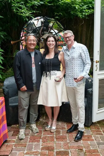 Three people posing for a photo in a garden setting. The man on the left wears a black jacket over a gray t-shirt and khaki pants. The woman in the center wears a black corset top and a cream-colored skirt. The man on the right wears a patterned blue and white shirt and light-colored pants.