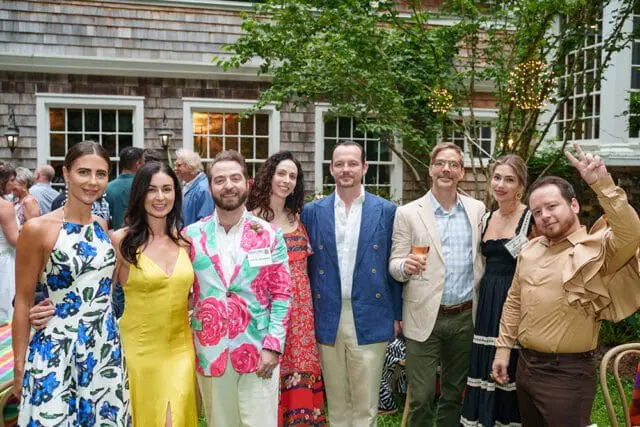 Group of people posing for a photo outdoors in front of a shingled building. A man in a floral jacket holds a name tag.