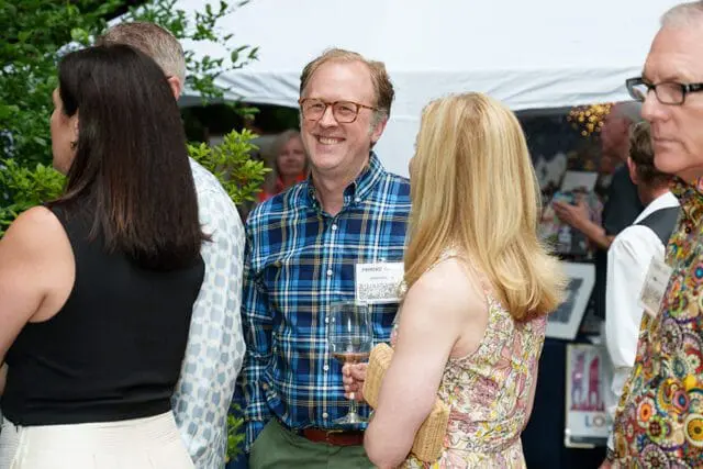 Man in blue plaid shirt smiles at a party, holding a wine glass. He wears glasses and has a name tag that reads 'PARADISE GARDEN'.
