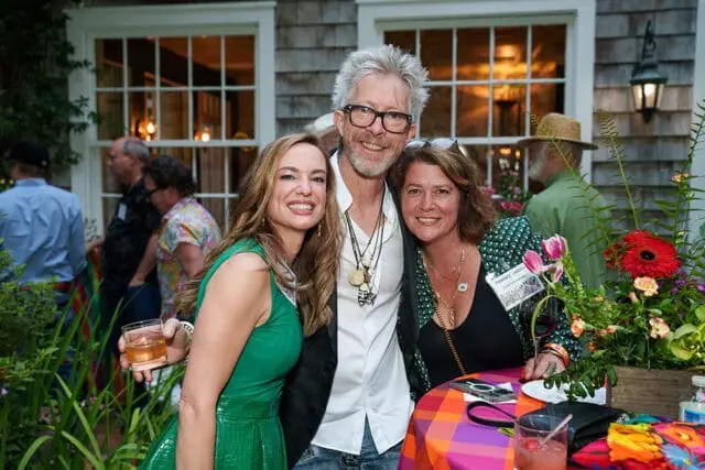 Three people smiling at a party. The woman on the left wears a green dress and holds a drink. The man in the center wears glasses and a white shirt. The woman on the right wears a patterned jacket and has a name tag that reads "PARADISE GARDEN".