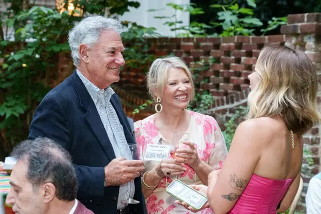 Man in a dark blazer and striped shirt smiles while holding a wine glass. A woman in a pink and white floral dress laughs, holding a wine glass and a phone.
