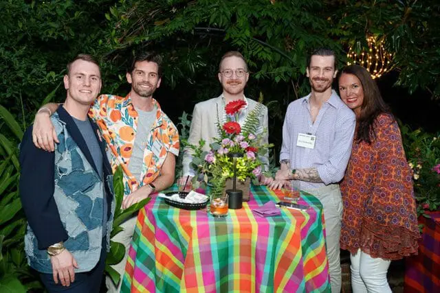 Five people pose around a table covered in a colorful plaid tablecloth at an outdoor event.