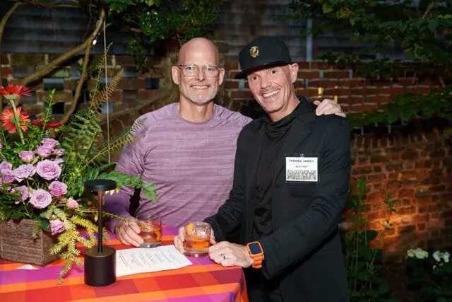 Two men smiling at a garden party. The man on the left wears glasses and a purple striped shirt. The man on the right wears a black jacket and hat, and has a "PARADISE GARDEN" name tag.