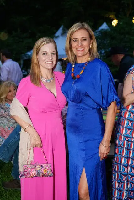 Two women smiling at an outdoor event. The woman on the left wears a bright pink jumpsuit and a light shawl, holding a colorful beaded clutch. The woman on the right wears a royal blue dress and a large beaded necklace.