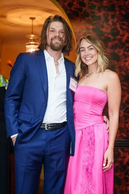 Man in a blue suit and white shirt stands next to a woman in a pink strapless dress with feathers.