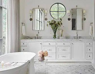 White bathroom with double vanity featuring marble countertops, brass sconces, and a round window. A freestanding bathtub is in the foreground.