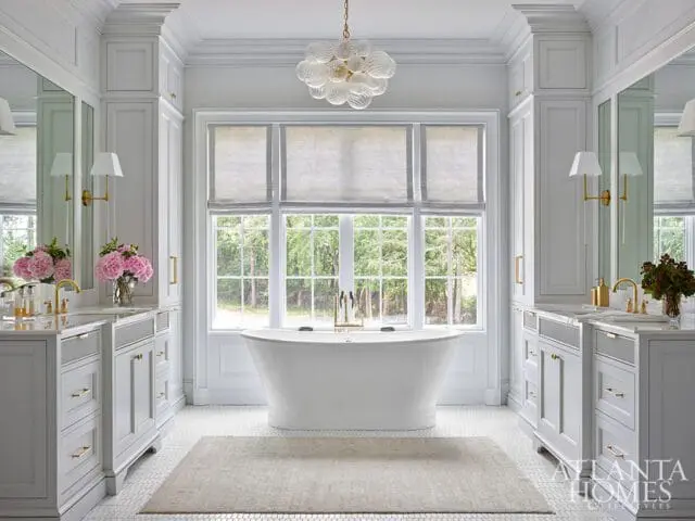 Luxurious white bathroom with a freestanding tub, dual vanities with marble countertops, and large windows overlooking a wooded area. Brass fixtures and a glass chandelier add elegant touches.