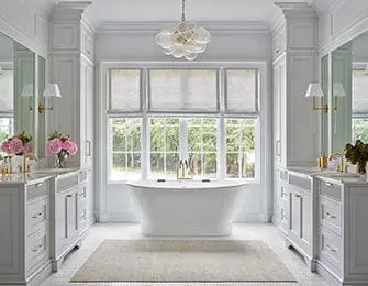 White bathroom with freestanding tub in front of a large window, flanked by double vanities with marble countertops and brass fixtures.